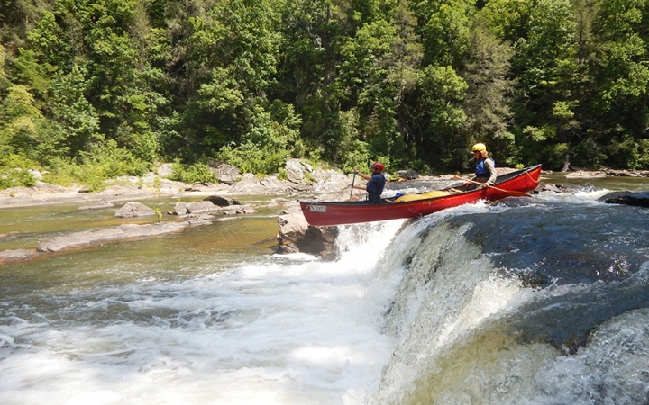 Two people wearing safety gear navigate a canoe over a short waterfall. Behind them is a rocky shore and dense trees.